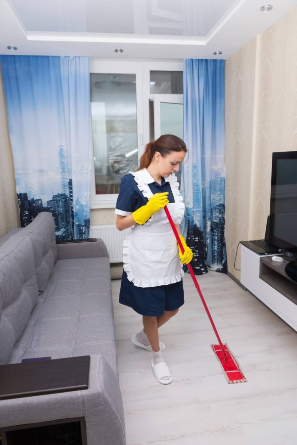 housekeeper-cleaning-hotel-suite-mopping-floor-comfortable-sofa-wall-mounted-television